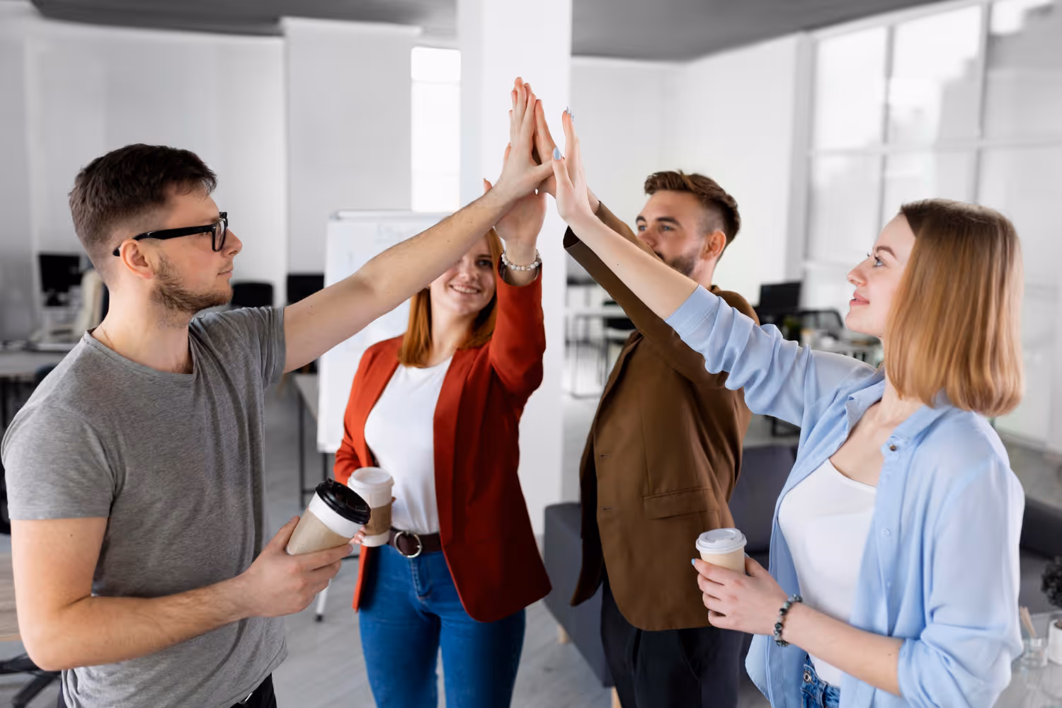 Four coworkers standing in office giving a group high-five while holding coffee cups.