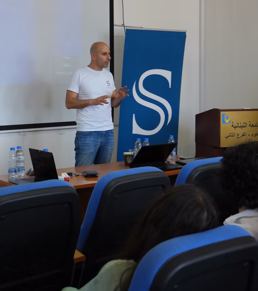 Man in white shirt giving a presentation in a classroom with audience seated in blue chairs.