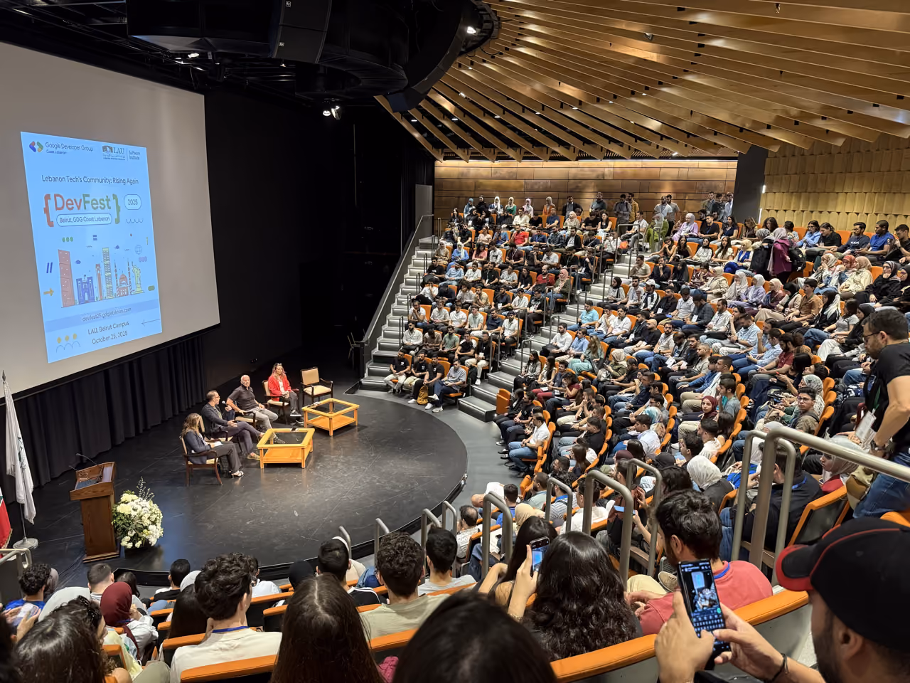 Man in white shirt giving a presentation in a classroom with audience seated in blue chairs.