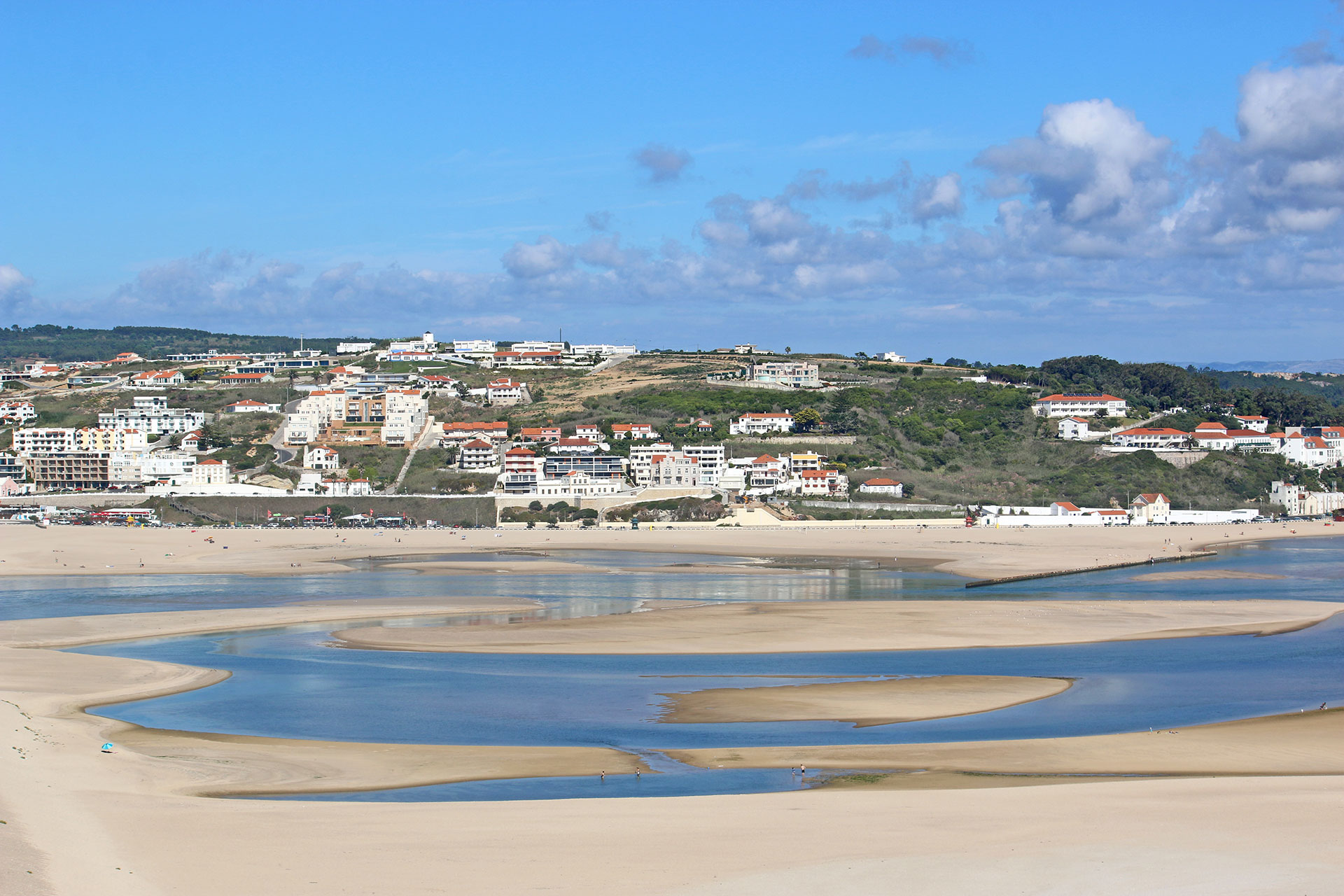 Bom Sucesso Beach curves around a tidal inlet, with hillside homes and golden sand under a bright blue sky