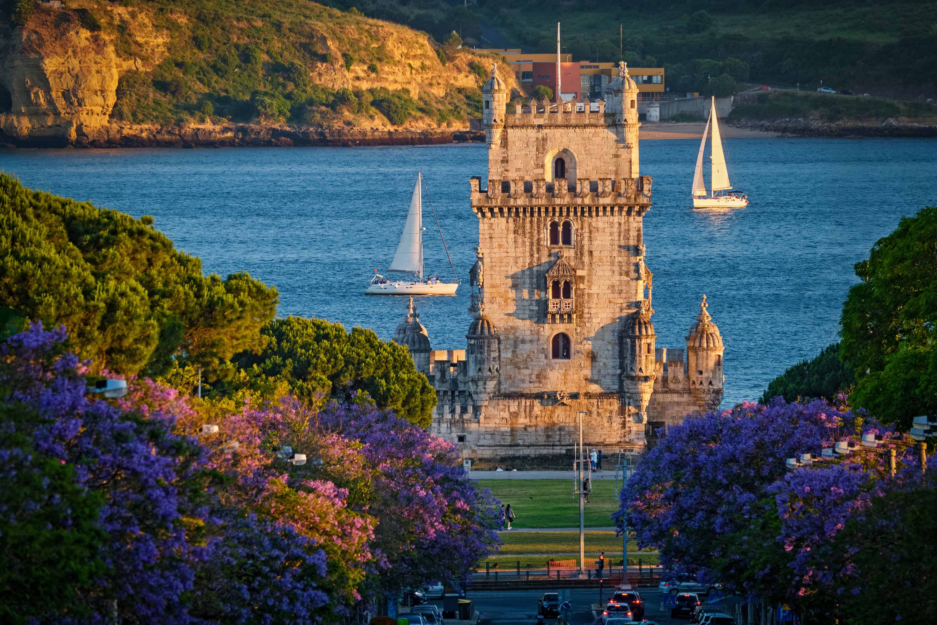 Scenic view of Belem Tower in Lisbon, Portugal, seen over a street with blooming purple jacaranda flower trees street with tourist sailboats on the Tagus River