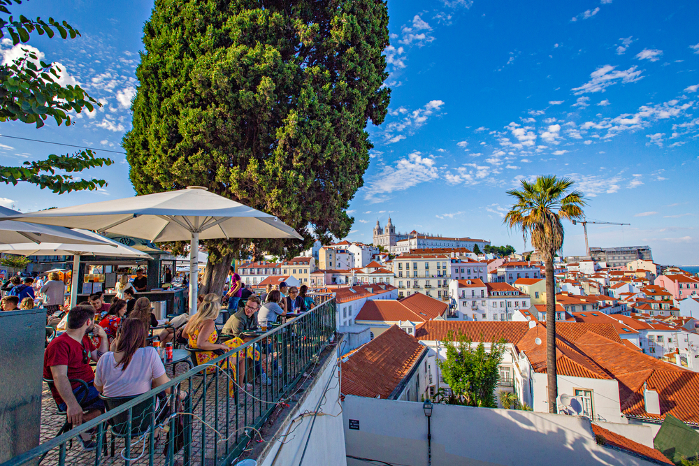 People enjoy drinks on a sunny terrace overlooking the red-tiled roofs of historic Lisbon
