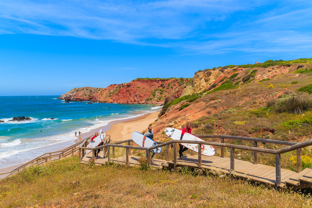 Surfers carry boards down a wooden staircase toward a sandy beach with colourful cliffs