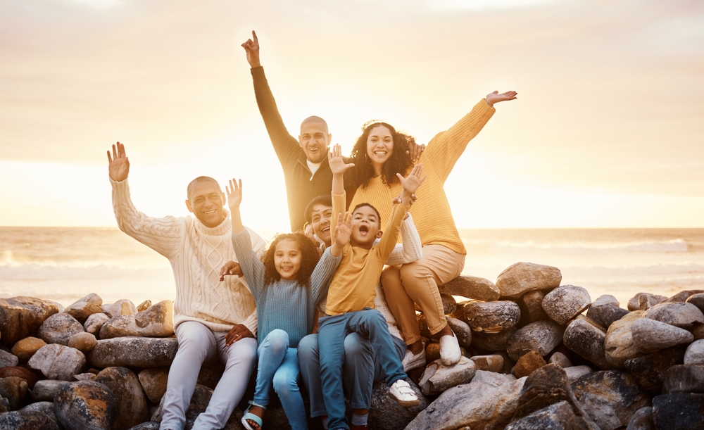 Happy multi-generational family sits on a rock wall by the ocean at sunset
