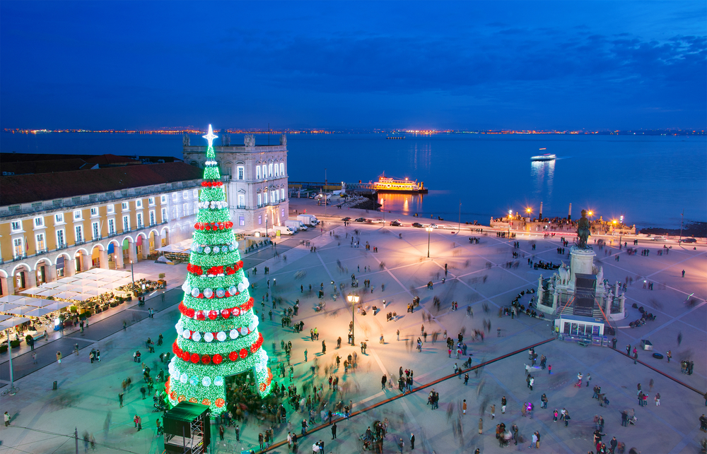 A towering, illuminated green Christmas tree stands in a bustling city square at dusk