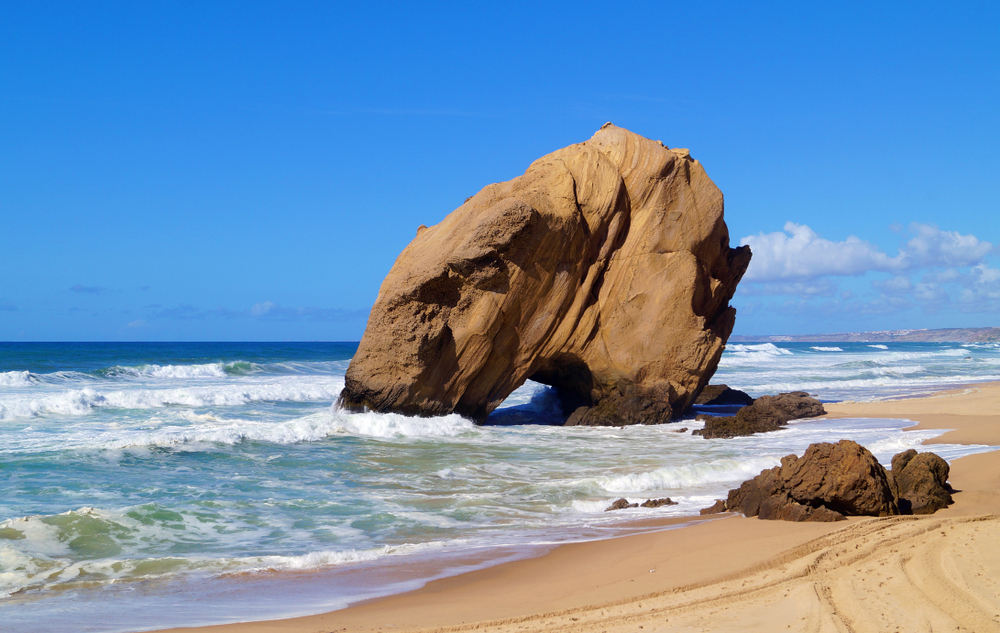 Huge tan boulder with a natural sea arch on a sandy beach as waves crash against the shore