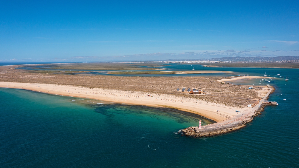 Aerial view of a thin sandy peninsula with a small red and white lighthouse at the tip