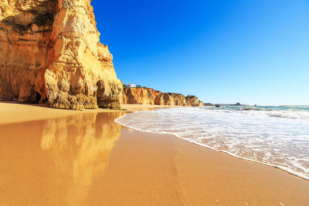 Golden sandstone cliffs reflected in the wet sand of a tranquil beach with blue water
