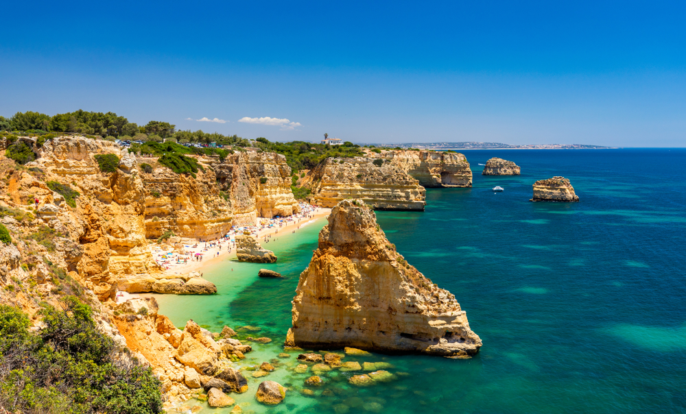 Golden sandstone cliffs and sea stacks surround a crowded beach with turquoise water