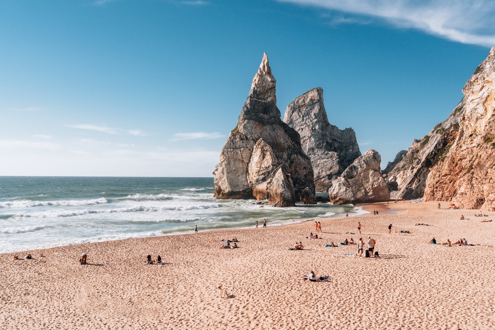 Beachgoers relax on the sand before a towering, pointed sea stack at Praia da Ursa