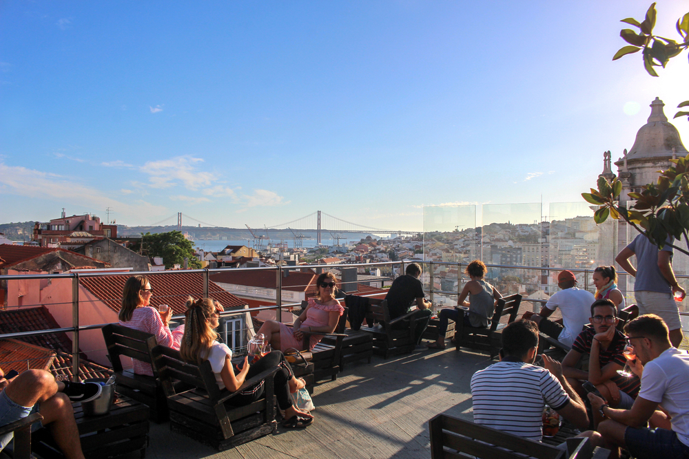 People enjoying drinks on a sunny Lisbon rooftop terrace with views of the April 25 Bridge