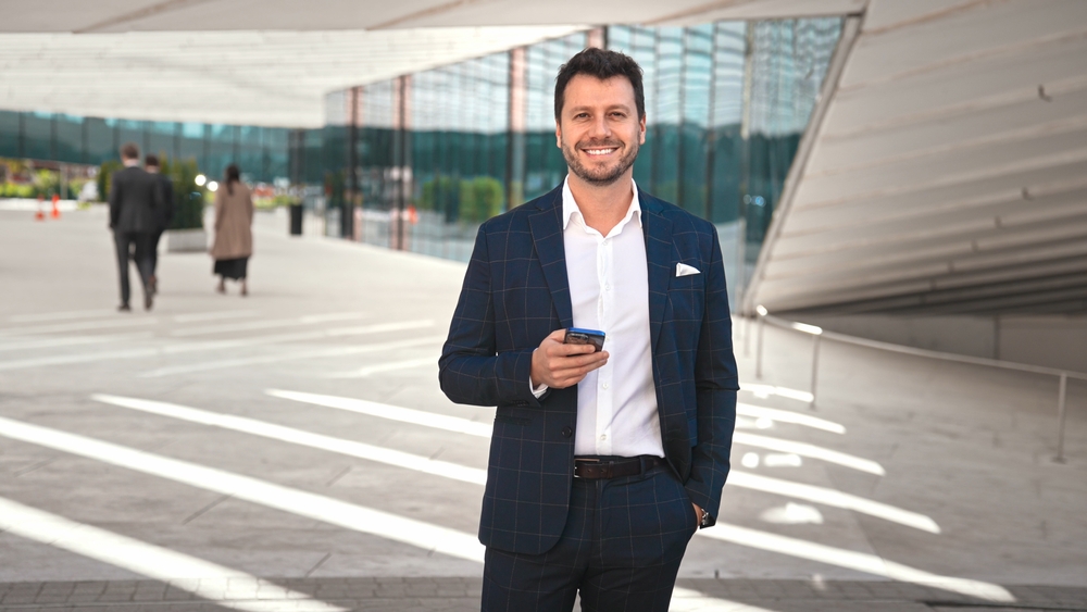 Confident man in tailored suit smiling with smartphone outside sleek, sunlit modern office building