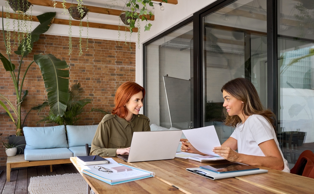 Two businesswomen working on a laptop and reviewing documents in a bright outdoor office