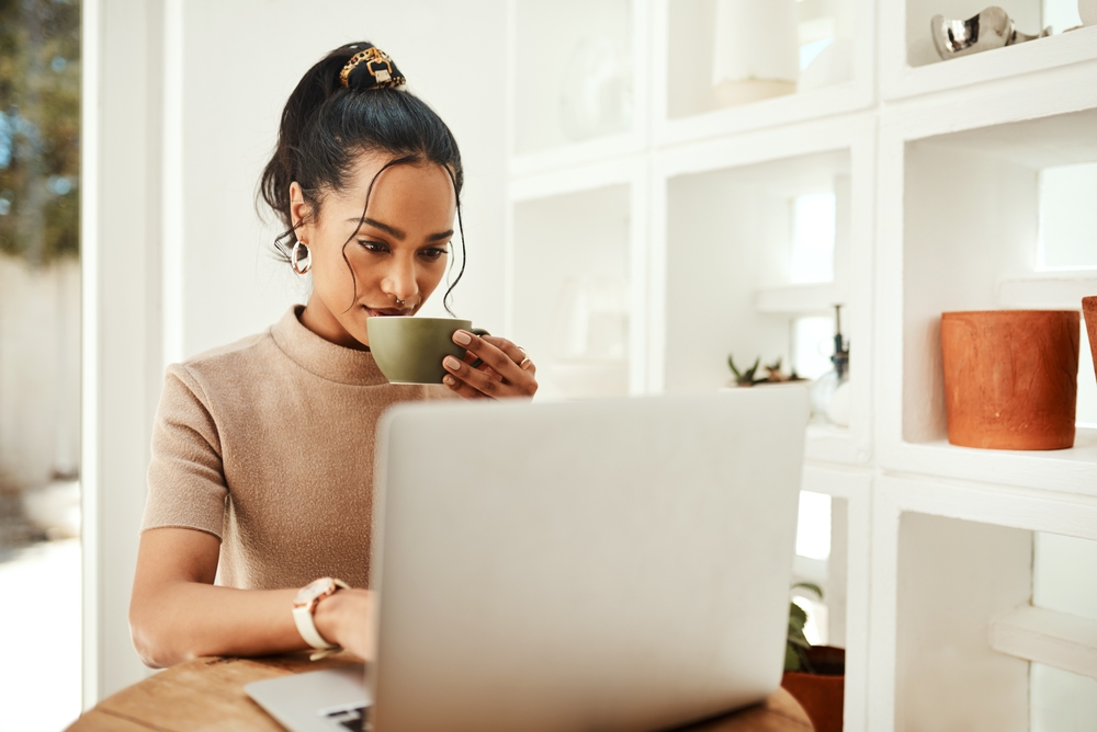 Businesswoman looking at a laptop and drinking coffee while sitting at a table at home