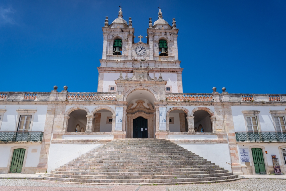 Grand white stone church with two bell towers and circular steps under a bright blue sky