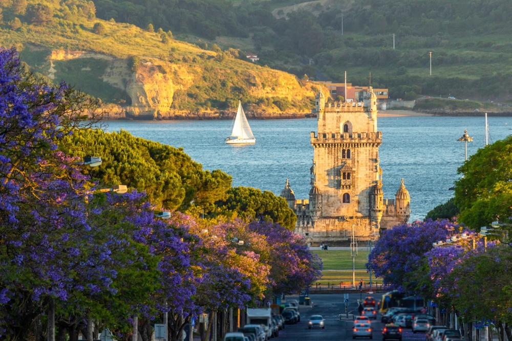Belem Tower stands at the end of a street lined with purple jacaranda trees in Lisbon