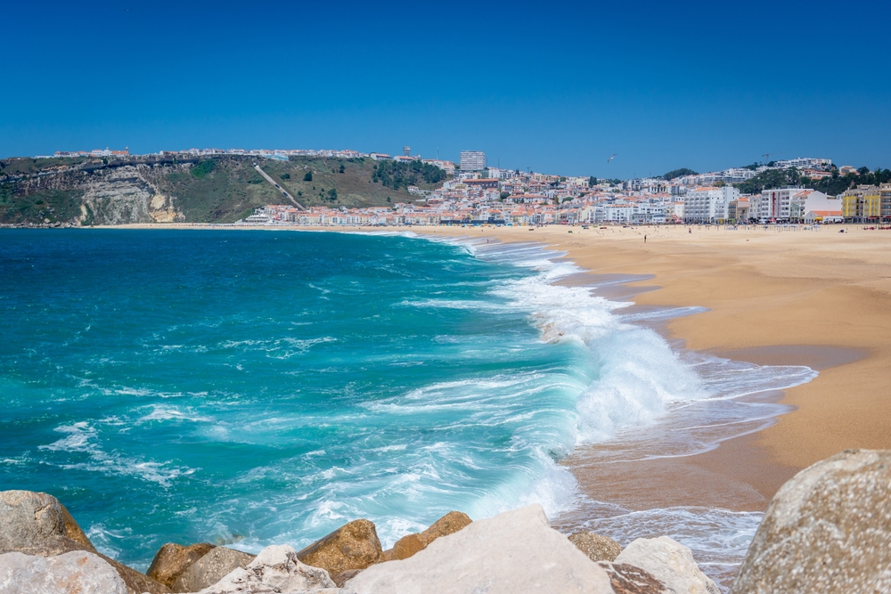 Turquoise waves crash on a golden sand beach below a coastal town and cliffs in Portugal