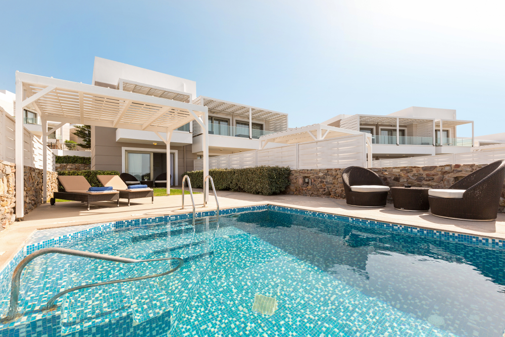 Sun loungers and a blue towel sit on a deck beside a clear pool at a modern white villa