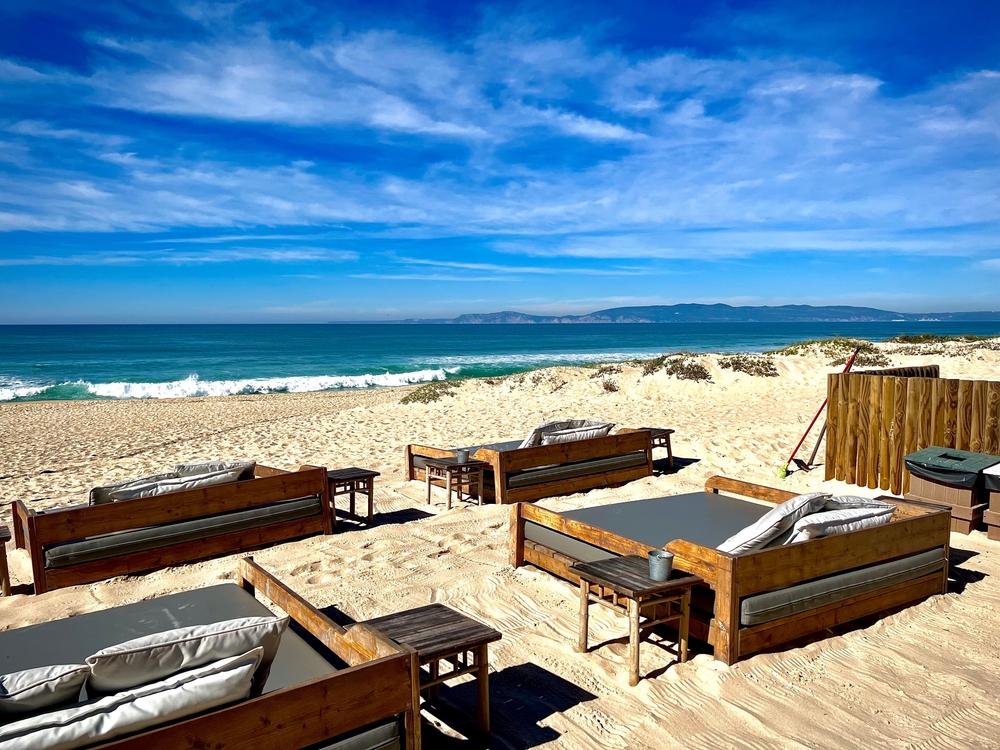 Beach lounge with wooden daybeds and grey cushions on a sunny sand beach by the blue ocean