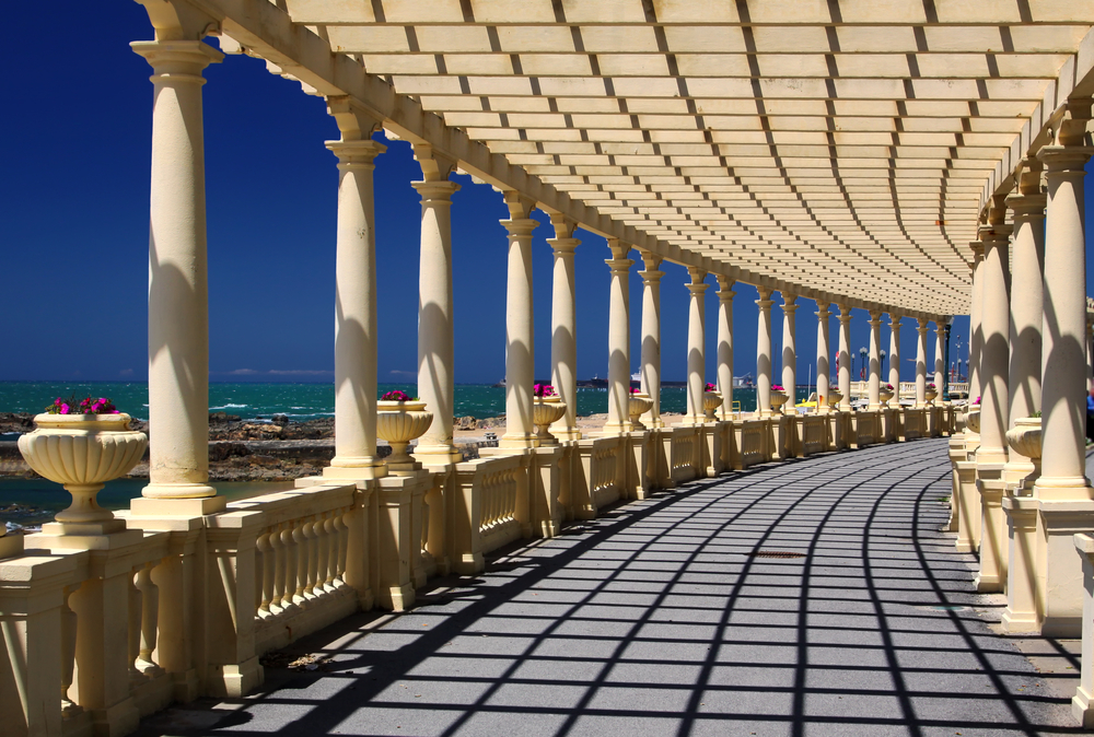 A scenic coastal promenade featuring a long curved pergola with white stone columns
