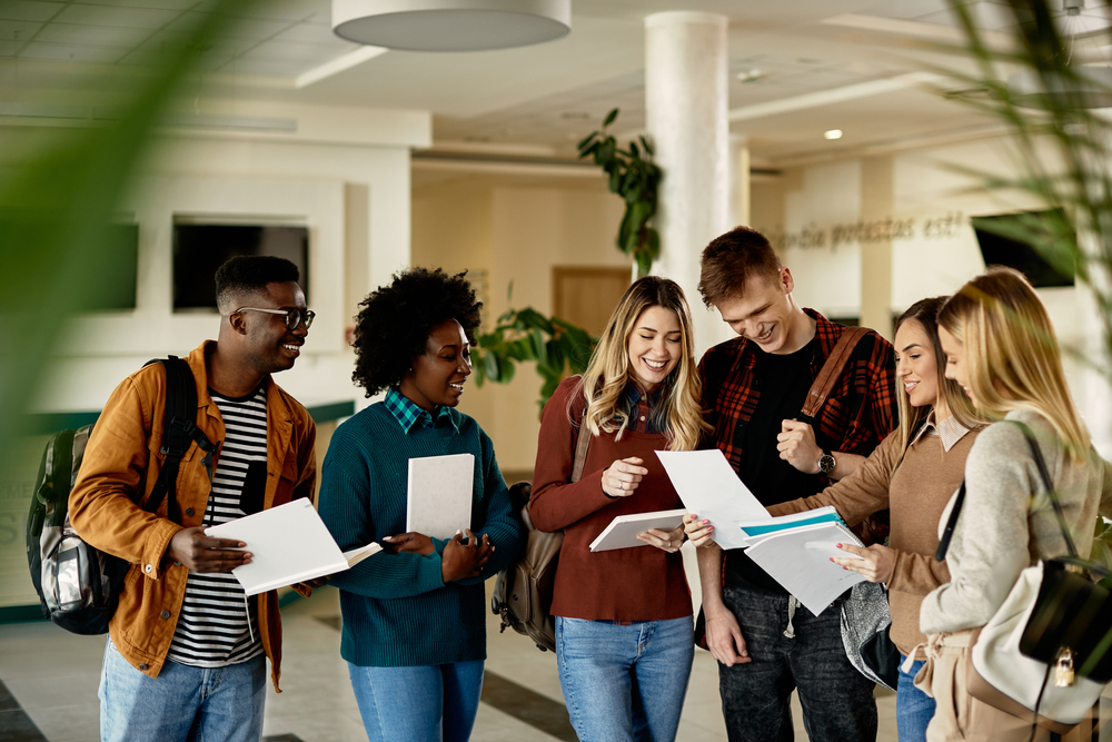 Group of diverse young students smiling and reviewing papers in a bright hallway