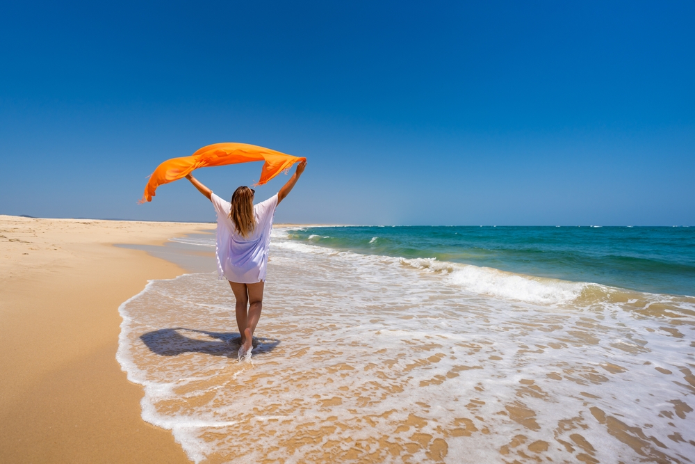 Woman in a white dress holding an orange scarf on a sunny beach with blue waves and sky