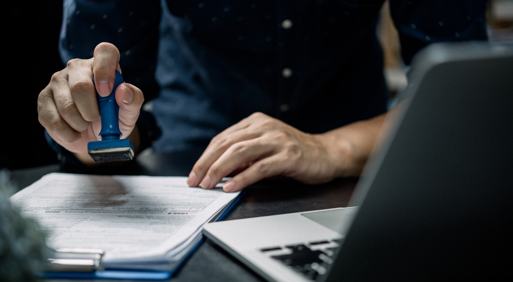 Hand pressing a blue rubber stamp onto an official document next to a silver laptop