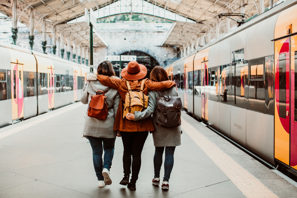 Three friends with backpacks walking arm-in-arm on a train station platform ready for travel