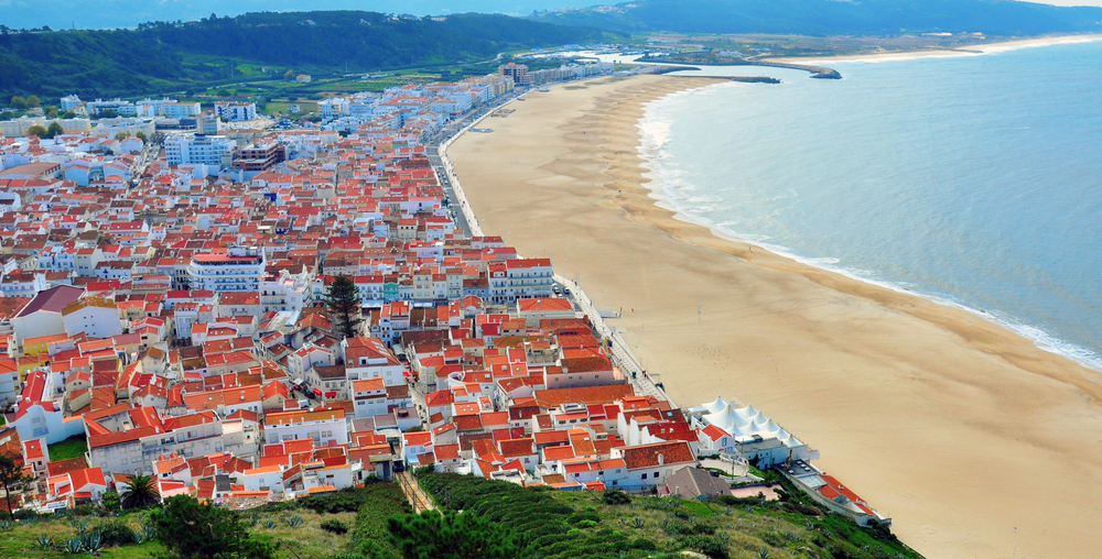 Aerial view of densely packed Nazaré with red-roofed homes bordering a wide sandy beach and ocean