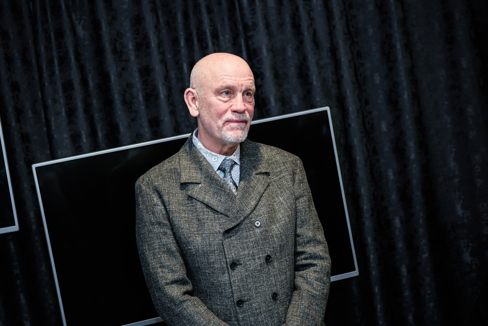 Actor John Malkovich in a gray double-breasted tweed coat speaking on stage
