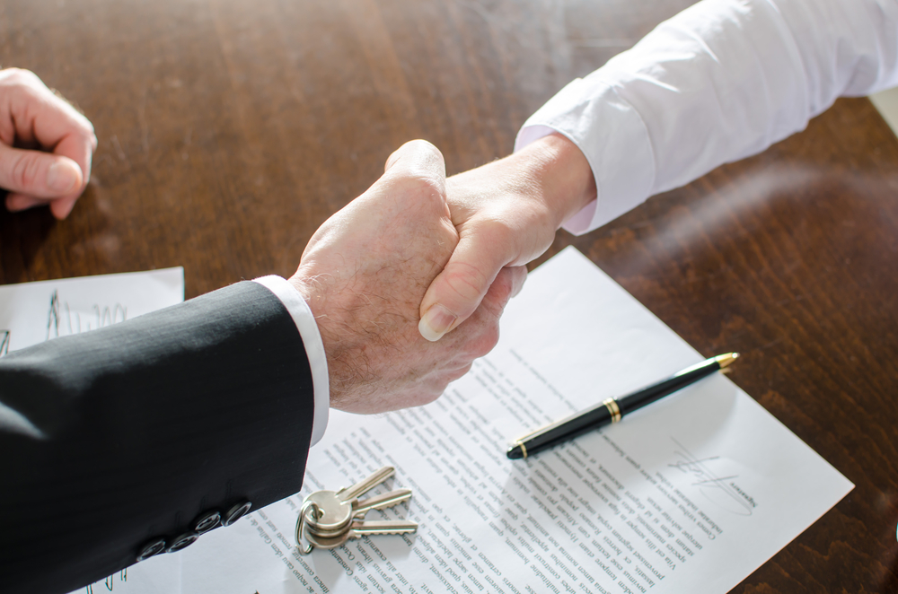 Business handshake agreement over signed contract documents and keys on an office desk