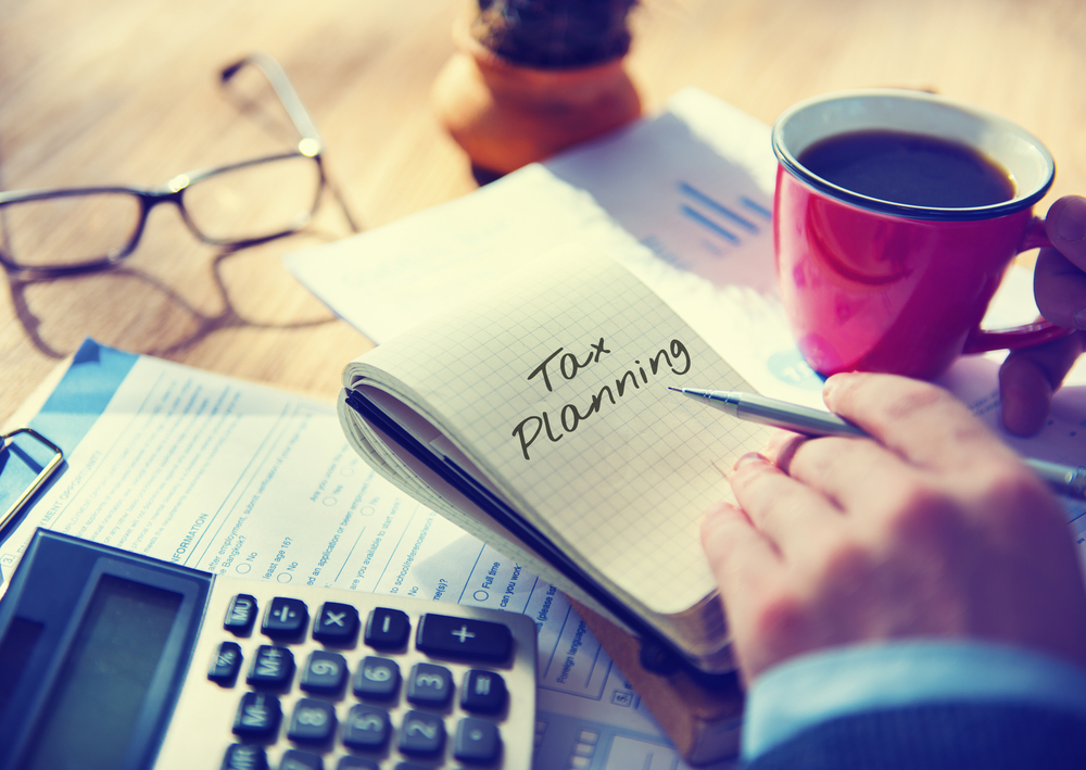 Person writing "Tax Planning" in a notebook with a calculator and coffee mug nearby