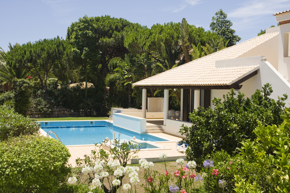 White Mediterranean villa with tan roof and blue swimming pool surrounded by lush garden