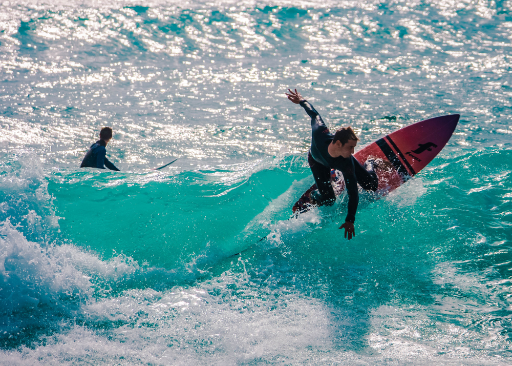 Surfer in black wetsuit rides vibrant turquoise wave while another surfer in water watches nearby