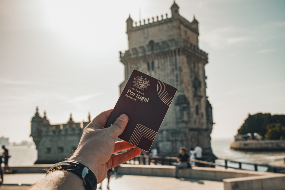 A hand holds a Portuguese passport with Belém Tower visible in the bright background