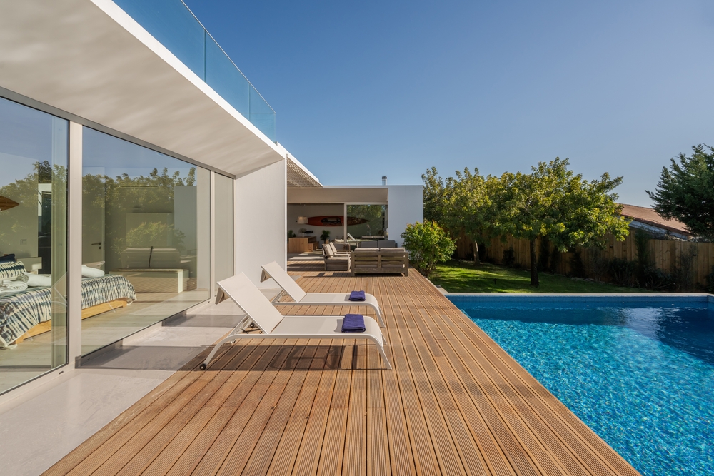 Modern villa with a blue swimming pool, wooden deck, and lounge chairs under a clear sky