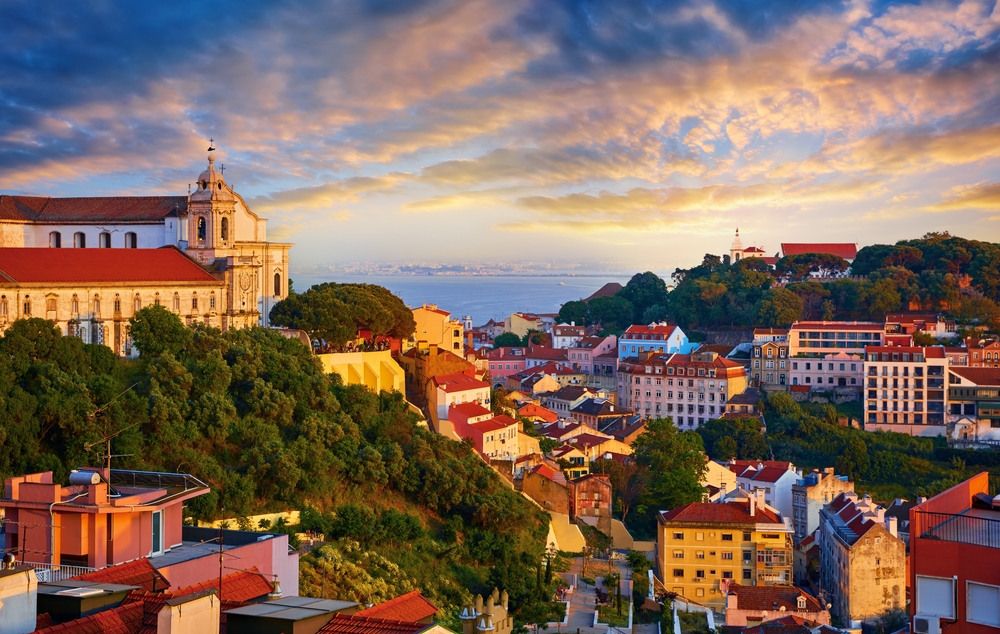 Sunset over Lisbon's Alfama district with red-roofed buildings and the Tagus River