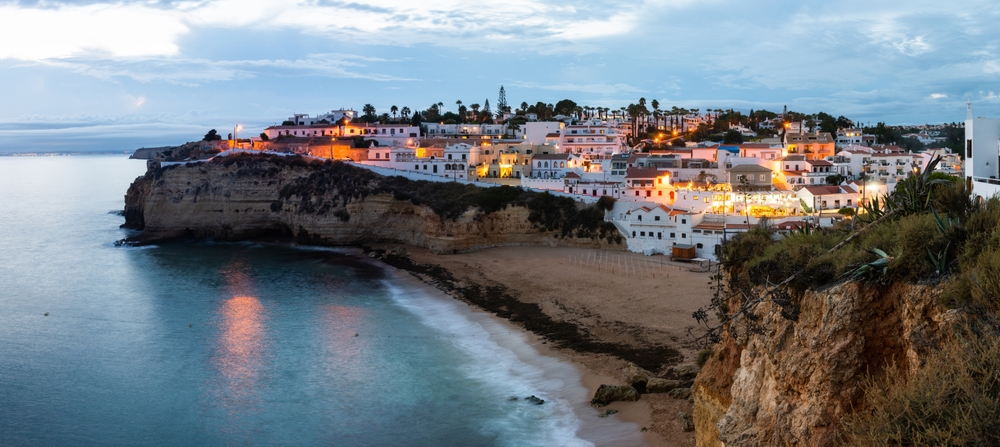 White houses with glowing lights perched on a dark cliffside overlooking a twilight beach
