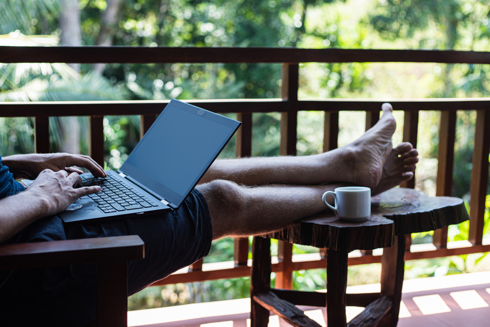 A person works on a laptop with their feet up on a balcony overlooking lush trees