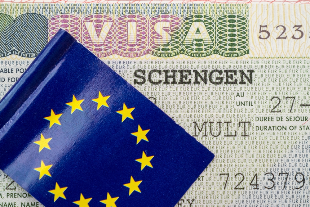 A blue passport cover with yellow stars partially hides a detailed Schengen visa page