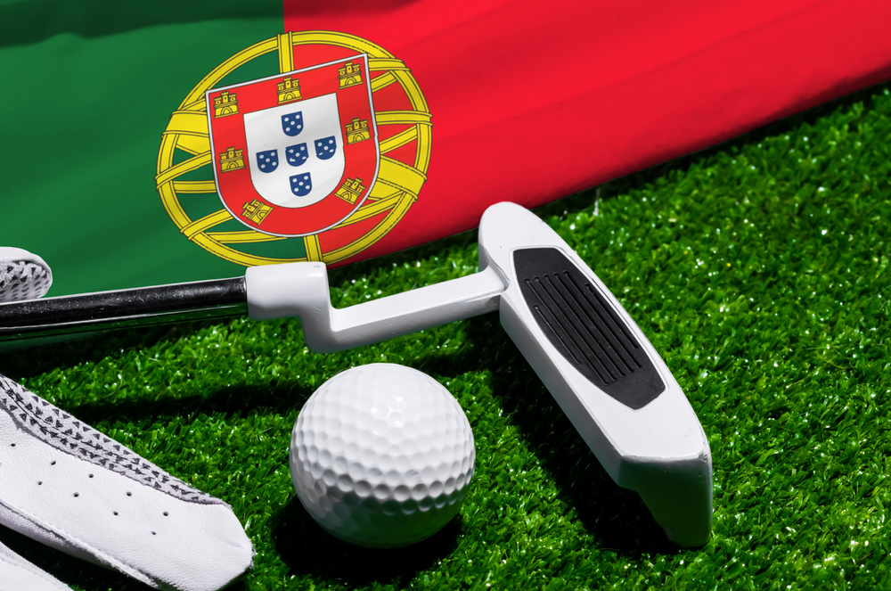 A white golf putter, ball, and glove rest on green turf in front of a Portuguese flag