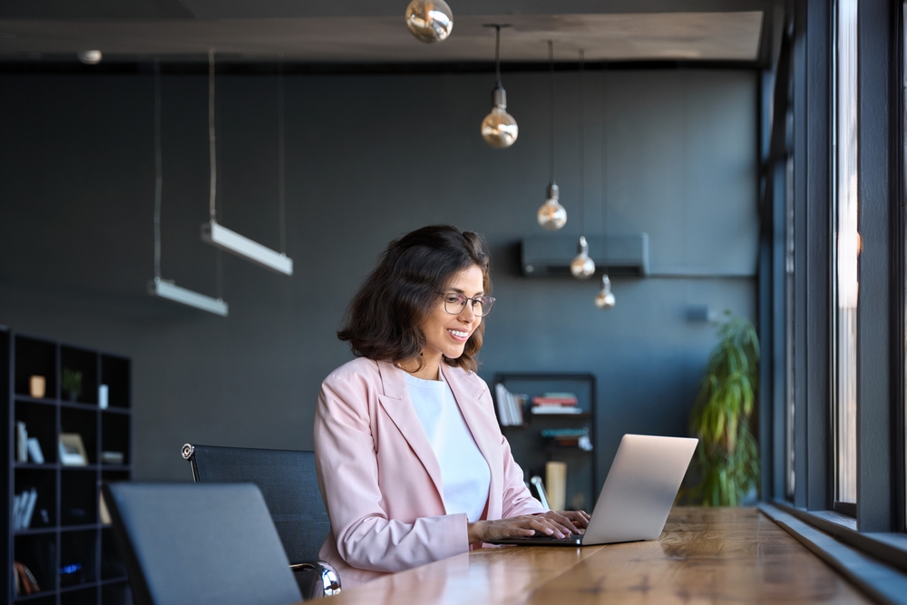 A woman in a pink blazer smiling while working on her laptop in a modern, sunlit office
