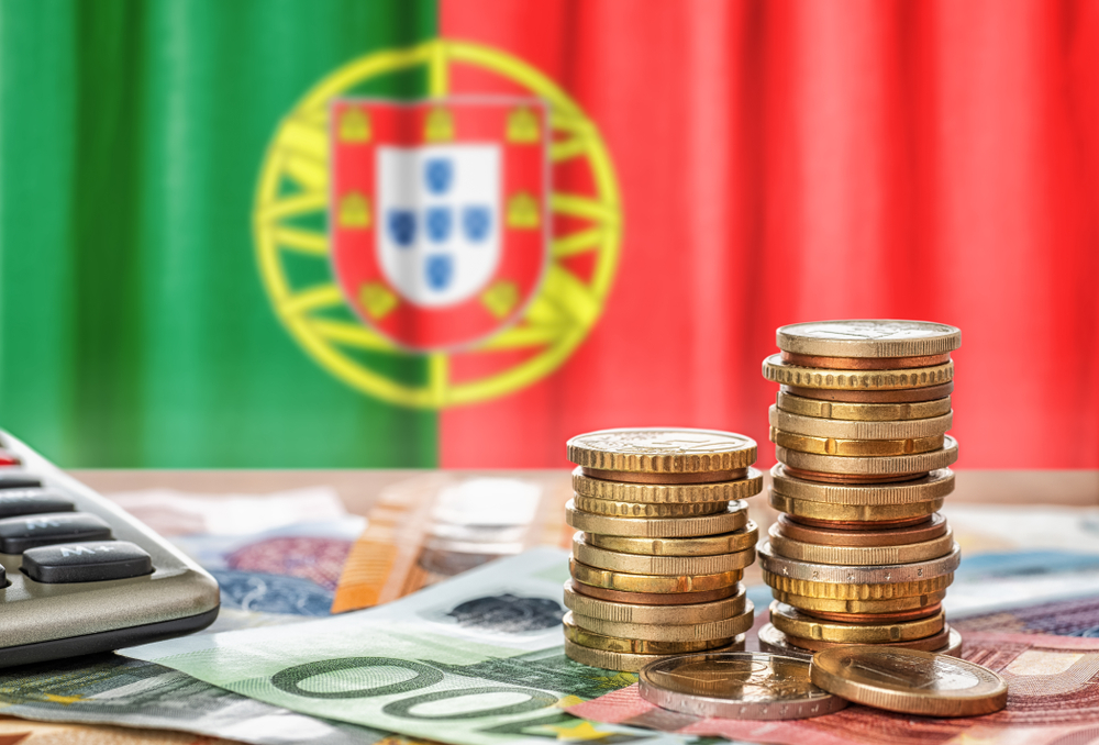 Stacks of Euro coins and banknotes on a desk, set against the backdrop of a Portuguese flag