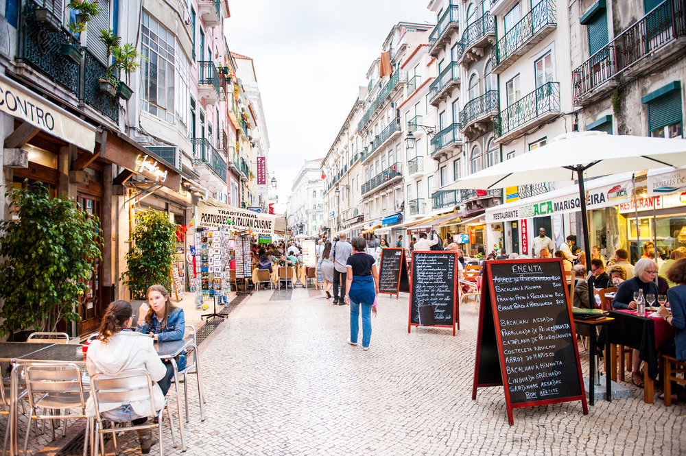 Busy pedestrian street in Lisbon with outdoor restaurant seating and chalkboard menus.