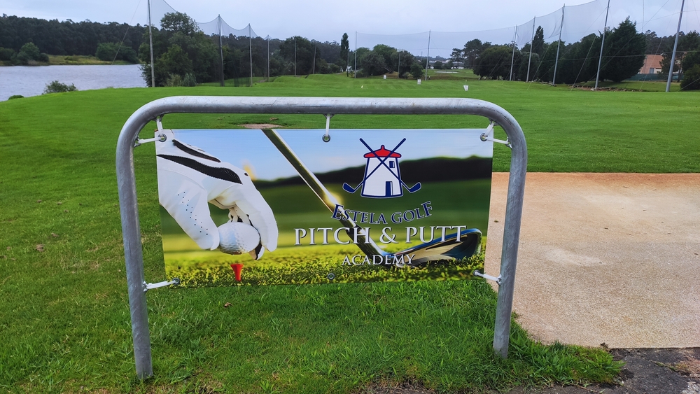 Sign for Estela Golf Pitch & Putt Academy in a green field next to a lake under a grey sky