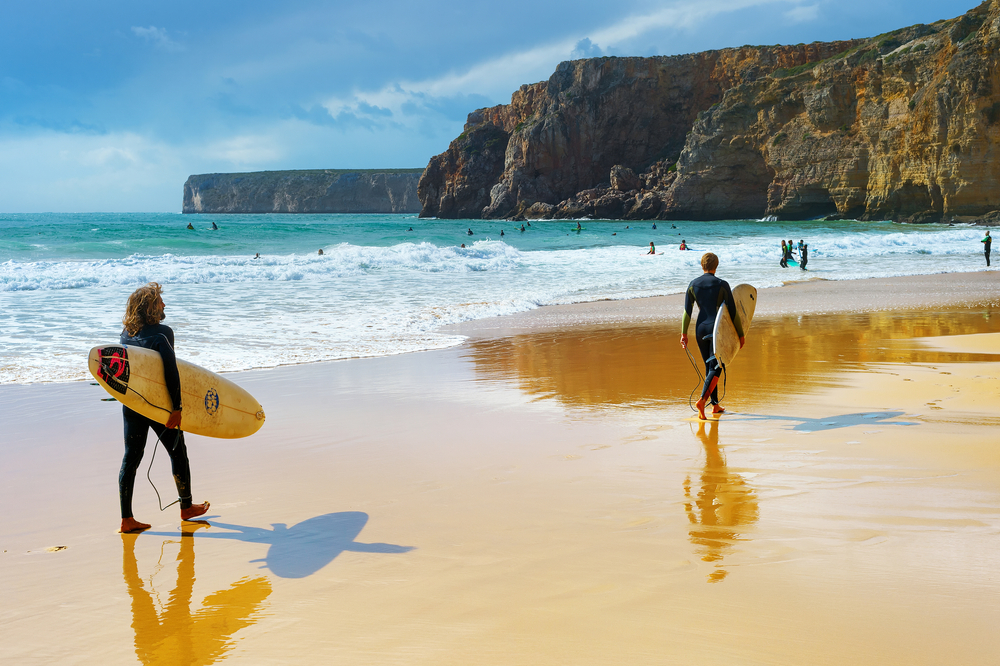 Two surfers carrying boards on a golden sandy beach with turquoise waves and rocky cliffs