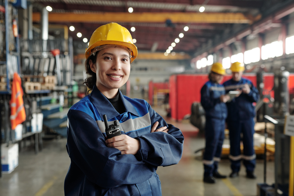 Smiling female engineer in a blue uniform and yellow hard hat holding a radio in a factory