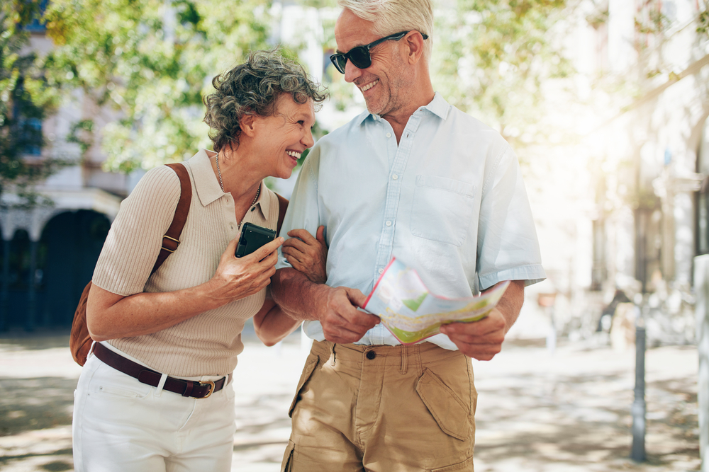 Happy older couple in sunglasses walking together and holding a map in a sunlit plaza