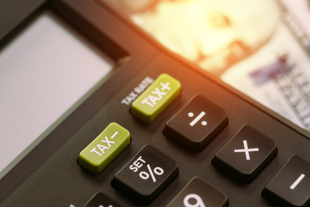 Close-up of a calculator with 'TAX+' and 'TAX-' buttons, near blurred money
