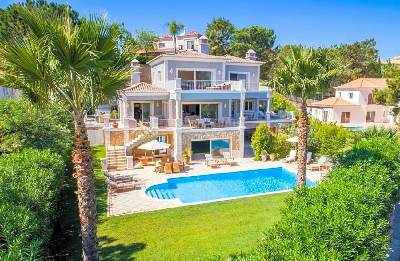 A three-storey luxury villa with a swimming pool and palm trees under a bright blue sky